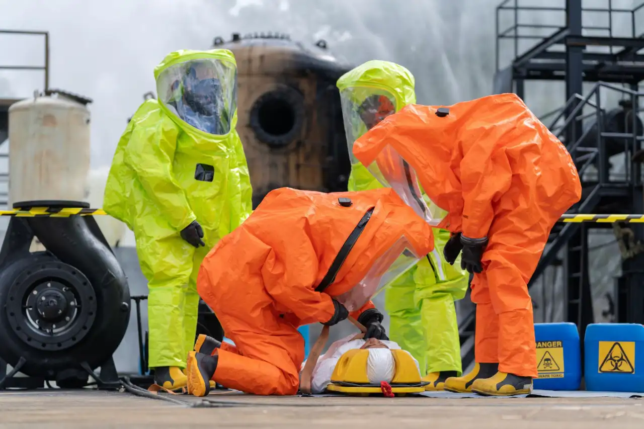 Team of worker, clad in protective gear including a gas mask and hazmat suit, rushes a patient on a field bed away from the hazardous area surrounding a chemical spill from a factory.